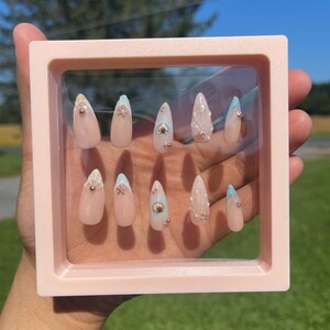 May include: A set of ten long, almond-shaped artificial nails displayed in a pink square frame. The nails are primarily nude with seashell and glitter accents. Some nails have a light blue tip.