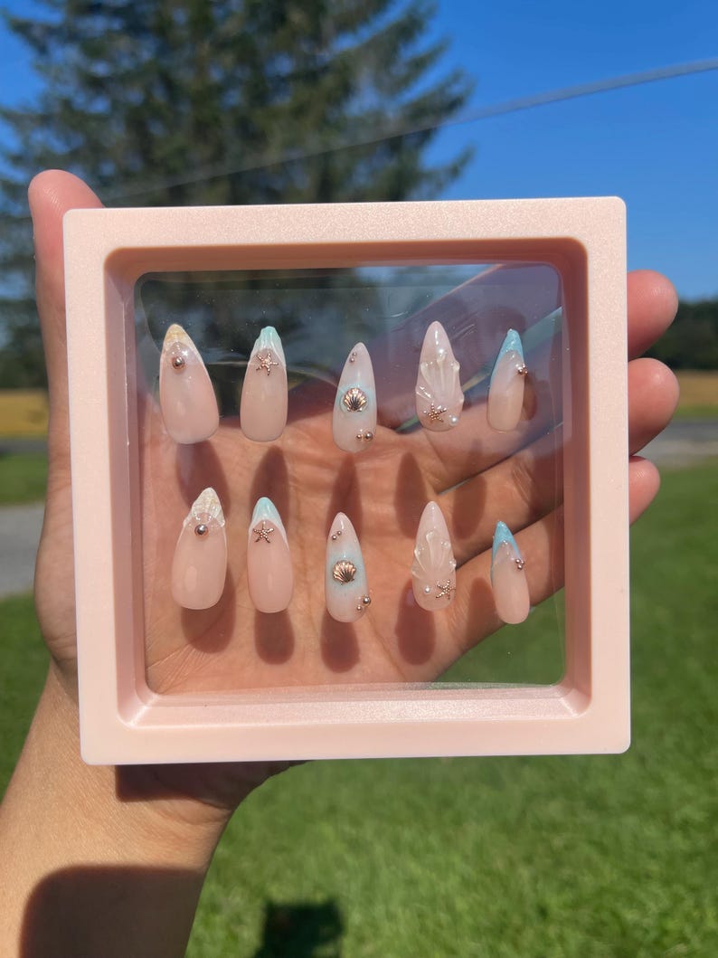 May include: A set of ten long, almond-shaped artificial nails displayed in a pink square frame. The nails are primarily nude with seashell and glitter accents. Some nails have a light blue tip.