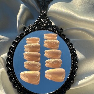 May include: A black, ornate hand mirror reflects a set of light peach-colored press-on nails. The nails are arranged in two columns of six, showcasing a glossy finish. The mirror's handle has a decorative floral design.