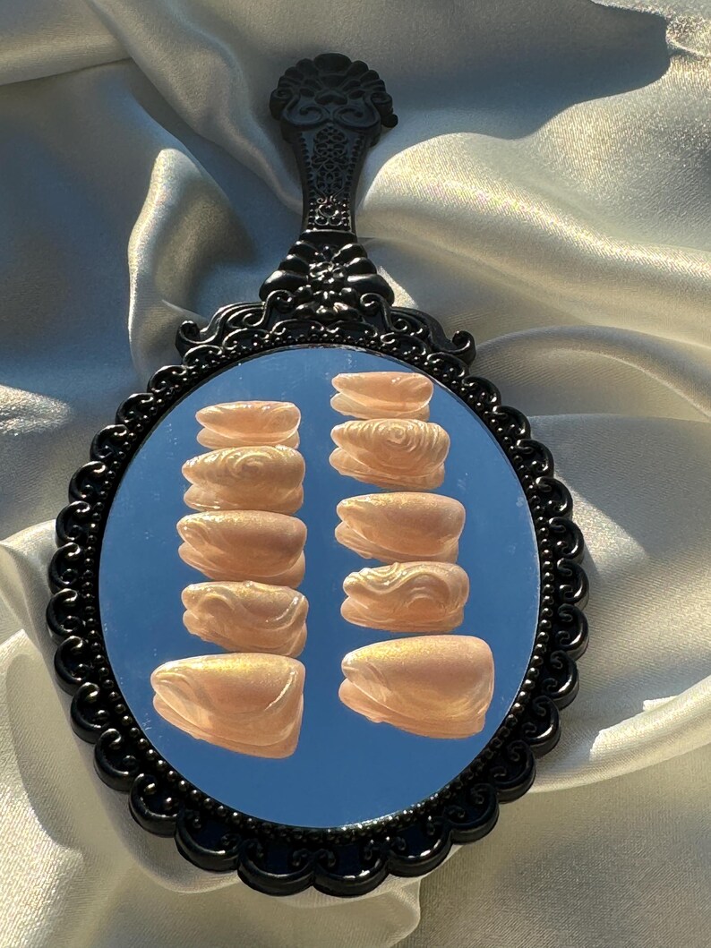 May include: A black, ornate hand mirror reflects a set of light peach-colored press-on nails. The nails are arranged in two columns of six, showcasing a glossy finish. The mirror's handle has a decorative floral design.
