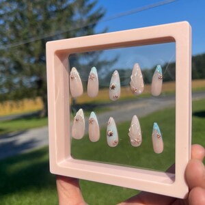 May include: A set of ten almond-shaped false nails in a pink frame. The nails feature a nude base with seashell and starfish embellishments. Some nails have a light blue tip. The frame is held up against a background of green grass and a blue sky.