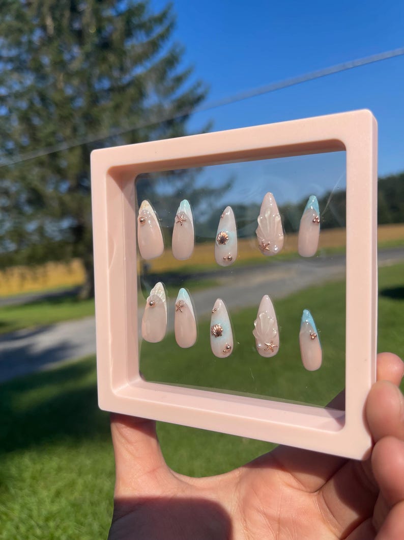 May include: A set of ten almond-shaped false nails in a pink frame. The nails feature a nude base with seashell and starfish embellishments. Some nails have a light blue tip. The frame is held up against a background of green grass and a blue sky.
