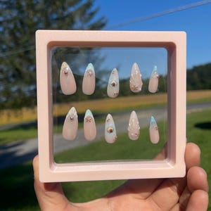 May include: A set of ten almond-shaped press-on nails in a pink frame. The nails are nude, with light blue and gold seashell and gem embellishments. The background shows a clear blue sky and green foliage.