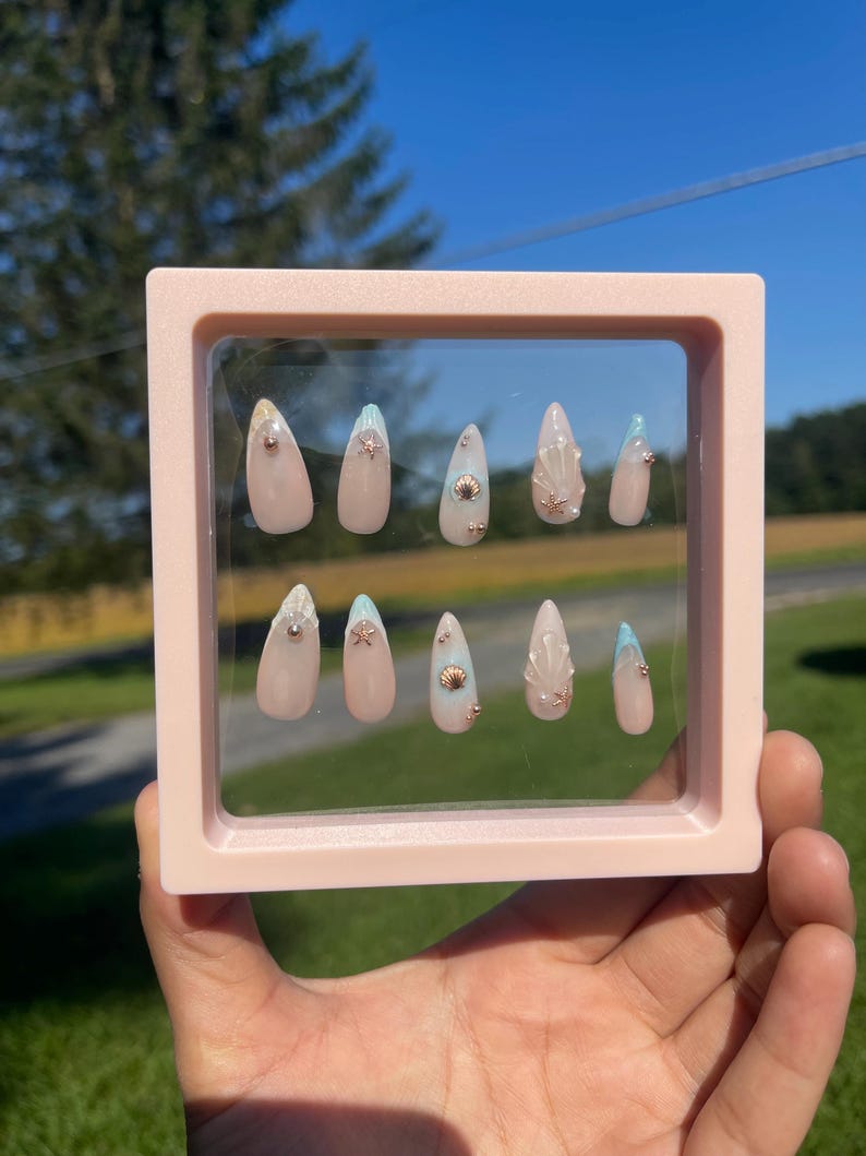 May include: A set of ten almond-shaped press-on nails in a pink frame. The nails are nude, with light blue and gold seashell and gem embellishments. The background shows a clear blue sky and green foliage.