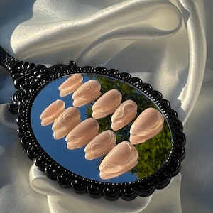 May include: A set of ten light peach-colored press-on nails, each with a sculpted design, are displayed on a black-handled mirror. The mirror reflects a green tree and blue sky. The mirror is set against a backdrop of white satin fabric.