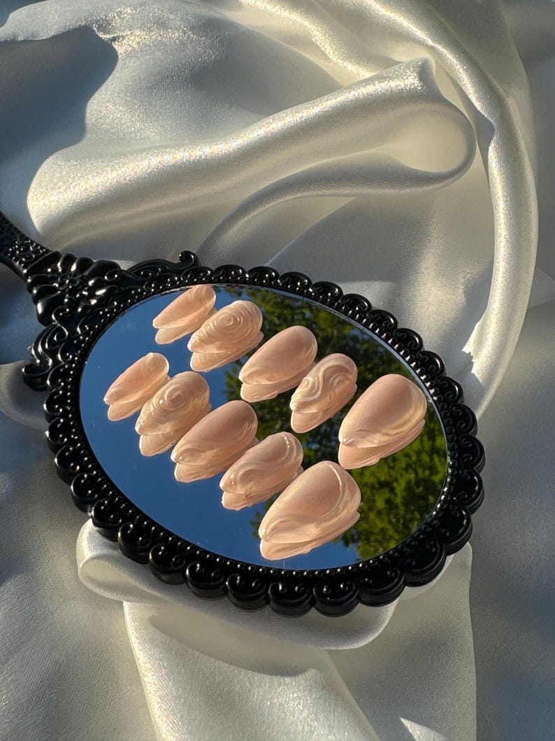 May include: A set of ten light peach-colored press-on nails, each with a sculpted design, are displayed on a black-handled mirror. The mirror reflects a green tree and blue sky. The mirror is set against a backdrop of white satin fabric.