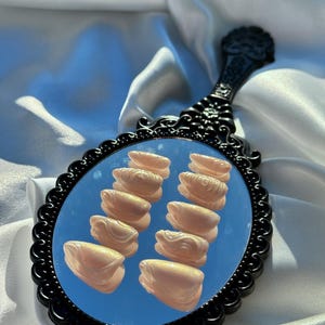 May include: A black, ornate hand mirror with a scalloped frame. The mirror reflects a blue sky and several light-colored, sculpted nail enhancements. The handle has a decorative floral design.