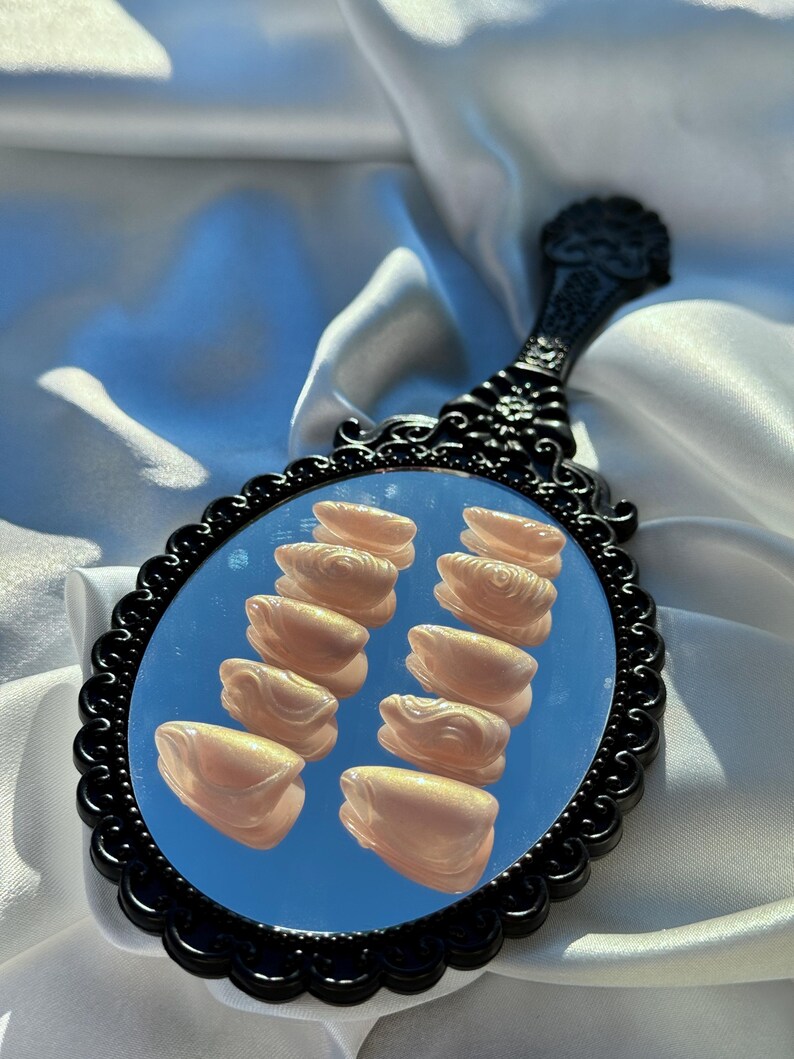 May include: A black, ornate hand mirror with a scalloped frame. The mirror reflects a blue sky and several light-colored, sculpted nail enhancements. The handle has a decorative floral design.