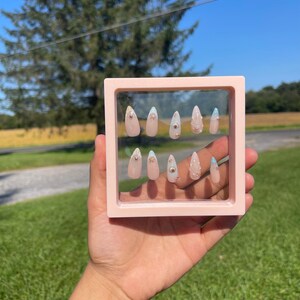 May include: A set of ten almond-shaped false nails presented in a pink square frame. The nails are a pale pink with gold studs and light blue accents. The background shows a sunny outdoor scene with green grass and a blue sky.