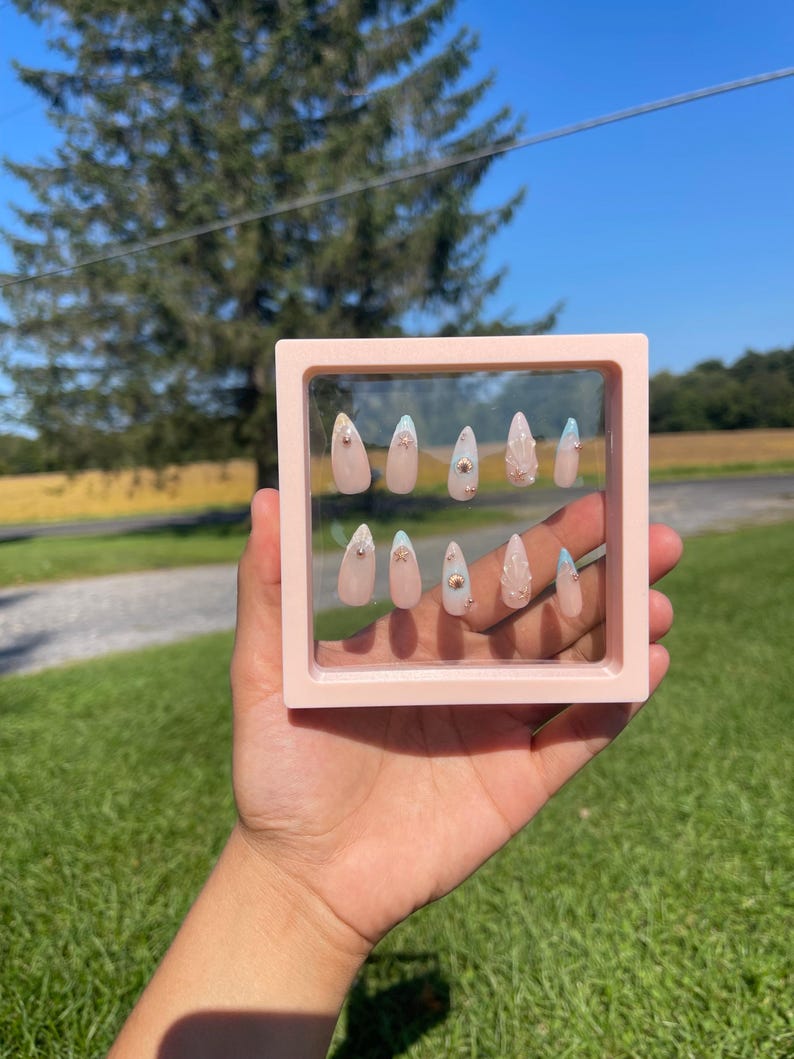 May include: A set of ten almond-shaped false nails presented in a pink square frame. The nails are a pale pink with gold studs and light blue accents. The background shows a sunny outdoor scene with green grass and a blue sky.