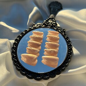 May include: A black-framed hand mirror with an ornate handle. The mirror reflects two stacks of light peach-colored, sculpted objects. The mirror is set against a backdrop of white fabric.