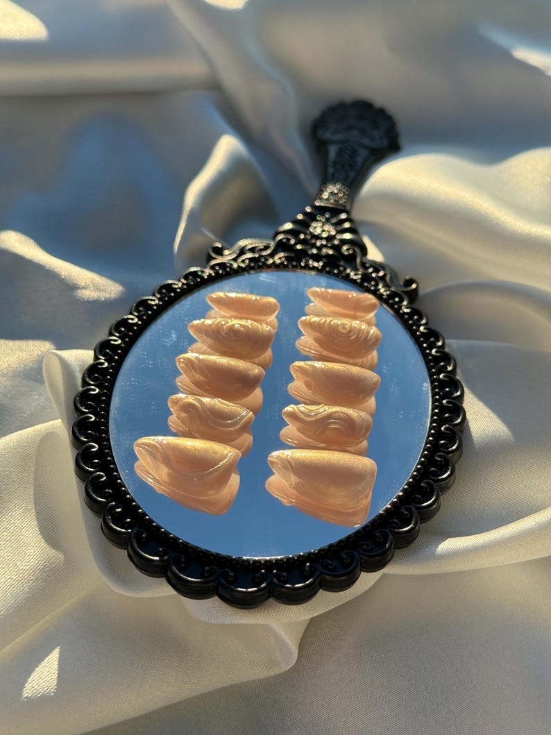 May include: A black-framed hand mirror with an ornate handle. The mirror reflects two stacks of light peach-colored, sculpted objects. The mirror is set against a backdrop of white fabric.
