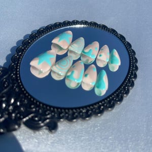 May include: A set of artificial nails displayed on a black framed mirror. The nails are almond-shaped with a light pink base and teal designs, including stars and swirls. The mirror has an ornate, decorative edge.