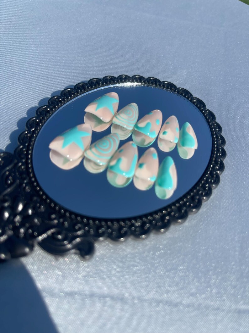 May include: A set of artificial nails displayed on a black framed mirror. The nails are almond-shaped with a light pink base and teal designs, including stars and swirls. The mirror has an ornate, decorative edge.