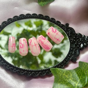 May include: A set of five pink artificial nails with a decorative pink design. The nails are rectangular and are arranged on a black framed mirror. Green leaves are in the background.