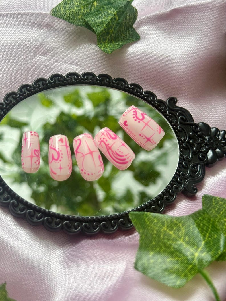 May include: A set of five pink artificial nails with a decorative pink design. The nails are rectangular and are arranged on a black framed mirror. Green leaves are in the background.