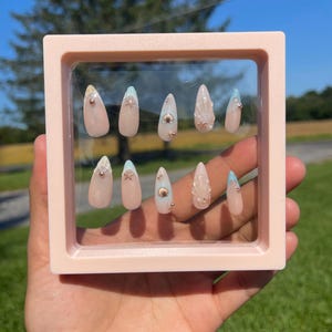May include: A collection of artificial nails presented in a pink square frame. The nails are pale pink with decorative elements such as gold beads, seashells, and light blue tips. The nails are almond-shaped.