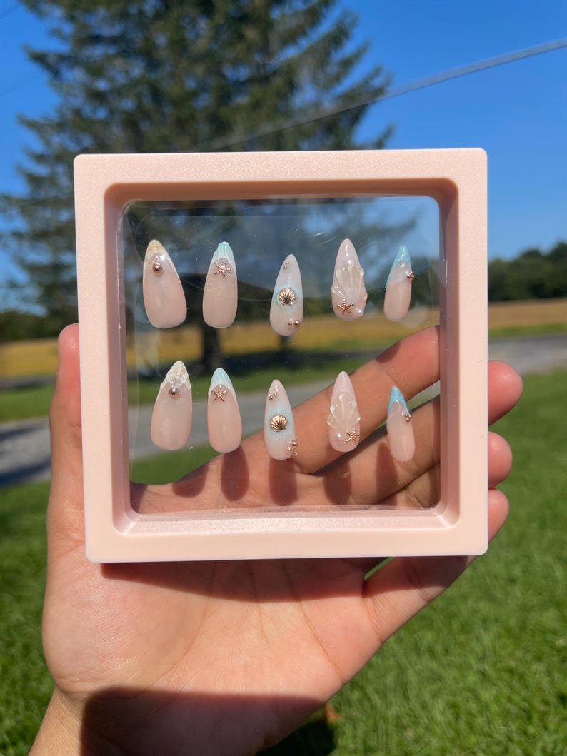 May include: A collection of artificial nails presented in a pink square frame. The nails are pale pink with decorative elements such as gold beads, seashells, and light blue tips. The nails are almond-shaped.