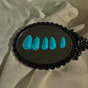 May include: A black, ornate mirror reflects six turquoise artificial nails. The nails are arranged in a row, showing different shapes. The mirror is set against a backdrop of cream-coloured, textured fabric.