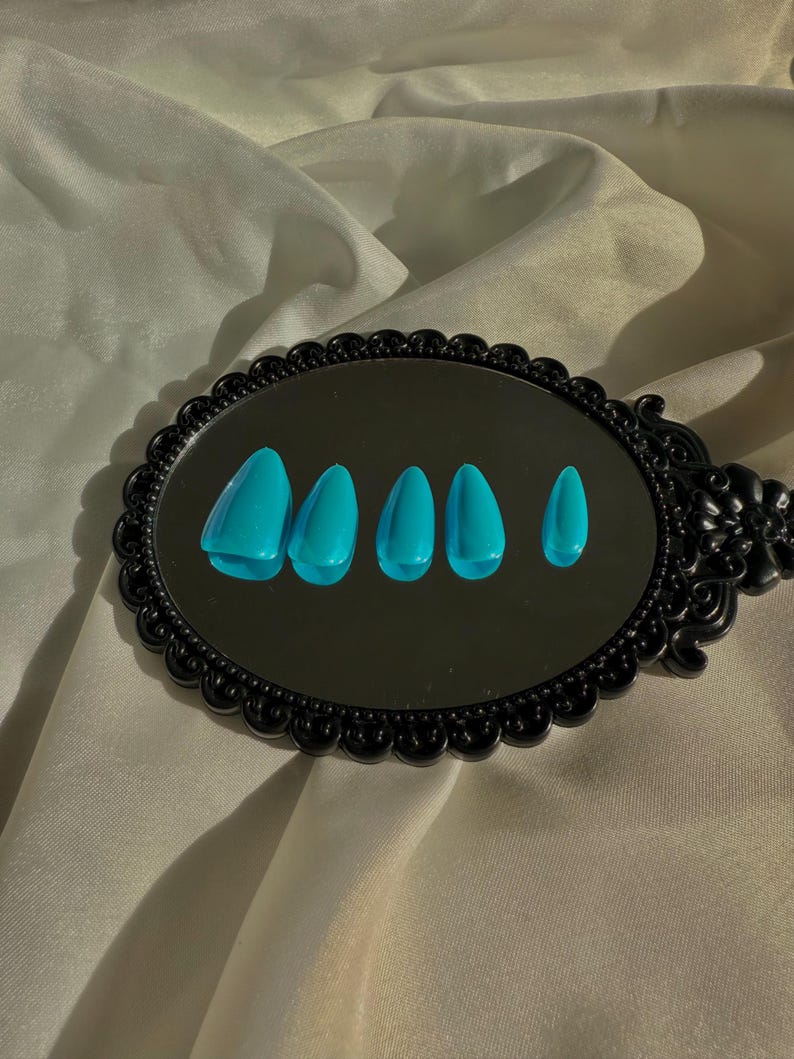 May include: A black, ornate mirror reflects six turquoise artificial nails. The nails are arranged in a row, showing different shapes. The mirror is set against a backdrop of cream-coloured, textured fabric.