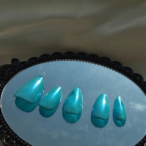 May include: Five teal-coloured, glossy, artificial nails are arranged on a black-framed oval mirror. The nails have a pointed shape and reflect light, creating a shimmering effect. The mirror is set against a neutral background.