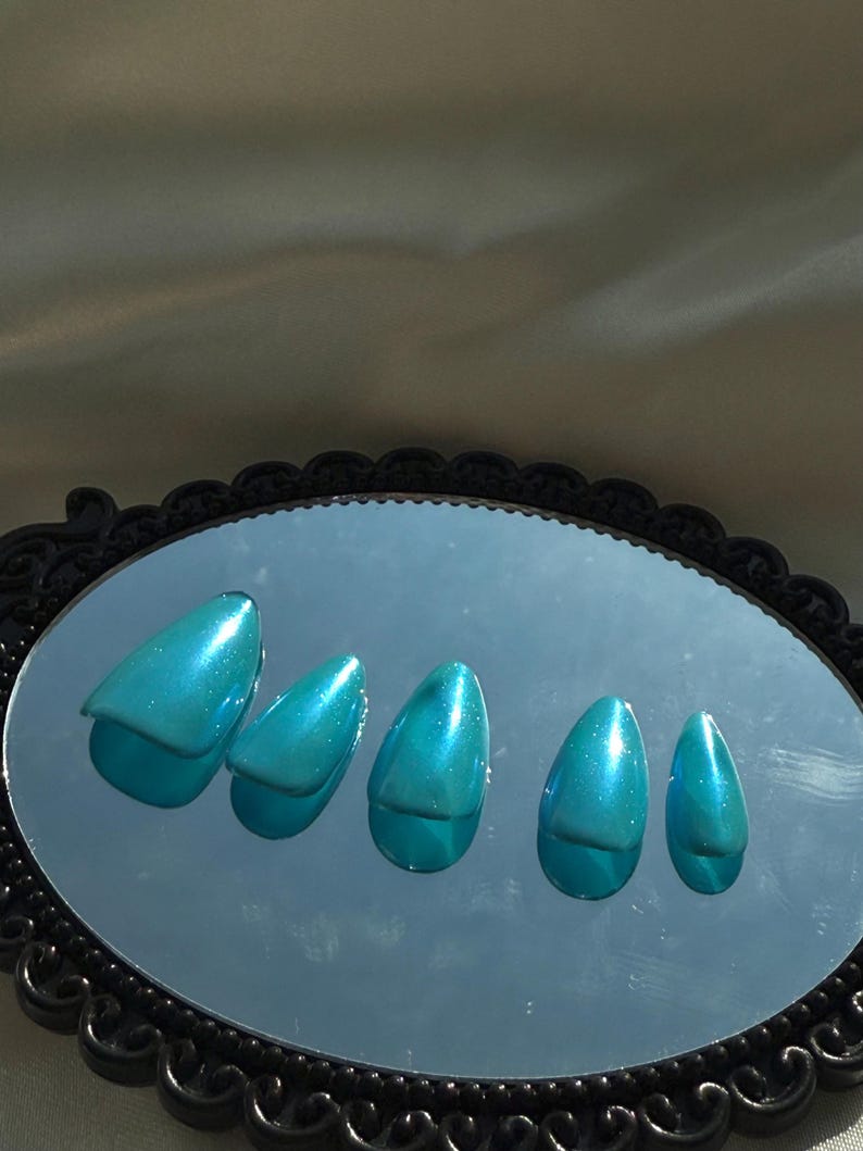 May include: Five teal-coloured, glossy, artificial nails are arranged on a black-framed oval mirror. The nails have a pointed shape and reflect light, creating a shimmering effect. The mirror is set against a neutral background.