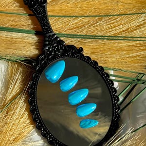 May include: A black, decorative hand mirror reflecting five bright blue, almond-shaped artificial nails. The nails have a glossy finish with subtle glitter and a marbled effect on the bottom nail. The mirror has an ornate handle.
