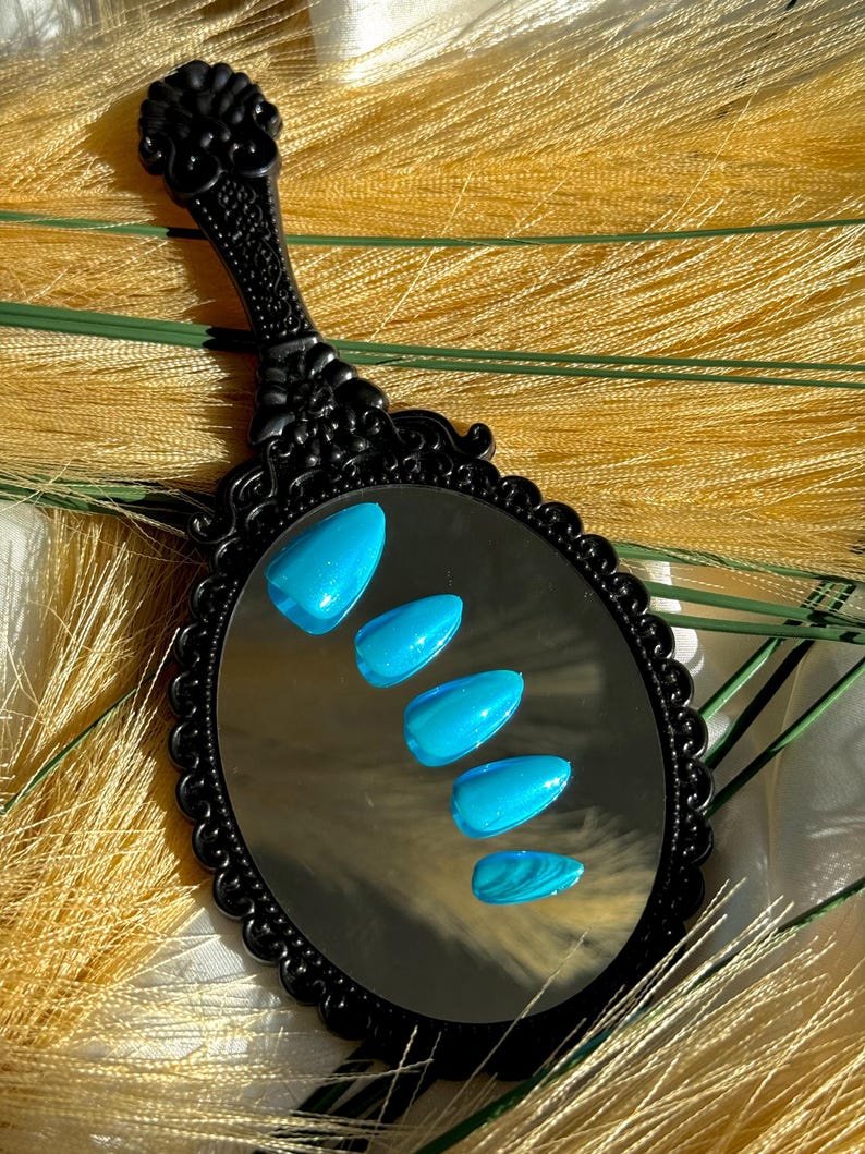 May include: A black, decorative hand mirror reflecting five bright blue, almond-shaped artificial nails. The nails have a glossy finish with subtle glitter and a marbled effect on the bottom nail. The mirror has an ornate handle.