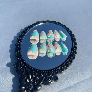 May include: A set of almond-shaped false nails with a light pink base and turquoise designs. The designs include starbursts, swirls, and French tips. The nails are displayed on a black, ornate mirror.