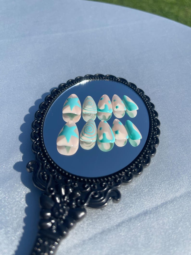 May include: A set of almond-shaped false nails with a light pink base and turquoise designs. The designs include starbursts, swirls, and French tips. The nails are displayed on a black, ornate mirror.