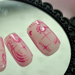May include: Close-up of artificial nails with a light pink base and a vibrant pink design. The design includes hearts, dots, and a grid pattern. The nails are displayed on a reflective surface.