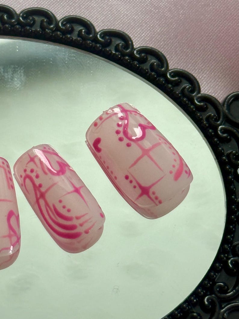 May include: Close-up of artificial nails with a light pink base and a vibrant pink design. The design includes hearts, dots, and a grid pattern. The nails are displayed on a reflective surface.