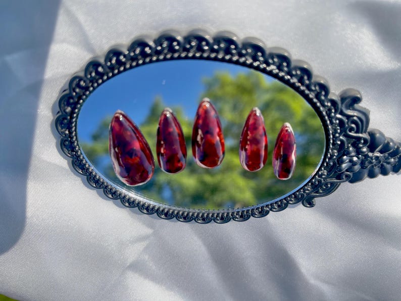 May include: Five almond-shaped, glossy, red and brown patterned artificial nails are displayed on a decorative, oval-shaped mirror with a black frame. The mirror reflects a blue sky and green foliage.