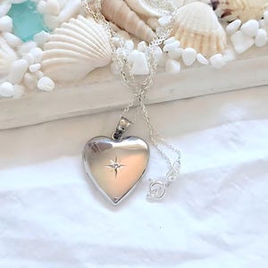 Puede incluir: Un colgante de relicario en forma de corazón de plata pulida suspendido de una delicada cadena de plata. El centro del corazón presenta una pequeña piedra clara incrustada con un grabado de estrella que irradia desde el engaste.