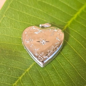 May include: A silver heart-shaped locket with a decorative floral and starburst design. The locket is set on a large green leaf. The locket has a small loop at the top for a chain. The heart is approximately 1.5 inches wide.