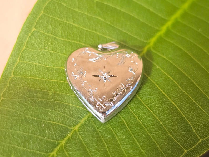 May include: A silver heart-shaped locket with a decorative floral and starburst design. The locket is set on a large green leaf. The locket has a small loop at the top for a chain. The heart is approximately 1.5 inches wide.