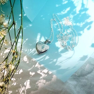 May include: A silver heart-shaped locket necklace with a delicate chain. The locket is detailed with an intricate design and rests on a light blue surface. White flowers and shadows add to the aesthetic. A beautiful piece of jewelry.