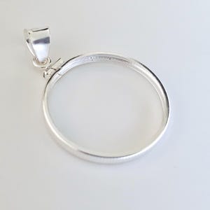 May include: A silver-colored pendant with a circular frame and a loop for a chain. The pendant has a bright, reflective surface and is set against a white background. The design is simple and elegant.