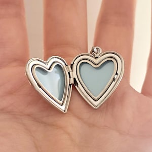 May include: Open silver heart-shaped locket with a light blue interior. The locket is open, revealing two heart-shaped spaces. The locket is held in an open hand. The locket has a small loop at the top for a chain.