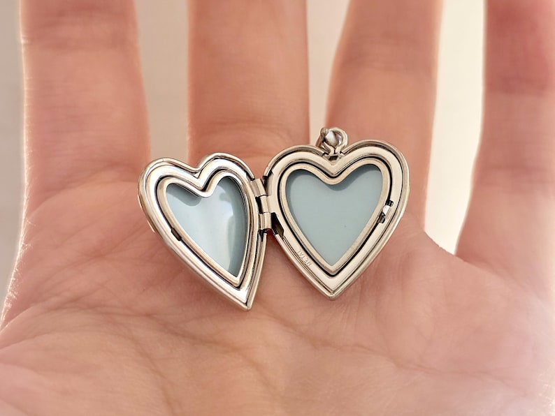 May include: Open silver heart-shaped locket with a light blue interior. The locket is open, revealing two heart-shaped spaces. The locket is held in an open hand. The locket has a small loop at the top for a chain.