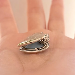 May include: A silver heart-shaped locket, open to reveal a light blue interior. The locket has a decorative floral design on the lid and a small loop for a chain. The locket is a piece of jewelry.