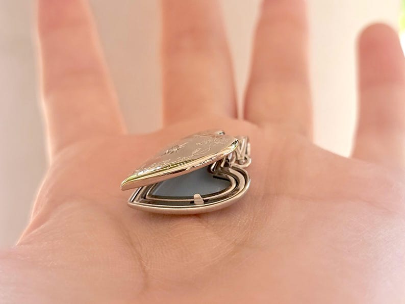 May include: A silver heart-shaped locket, open to reveal a light blue interior. The locket has a decorative floral design on the lid and a small loop for a chain. The locket is a piece of jewelry.