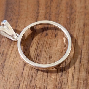 May include: A silver pendant with a circular design. The pendant has a loop at the top for a chain. The pendant is on a wooden surface.
