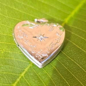 May include: A silver heart-shaped locket with a starburst design and floral accents. The locket is closed and sits on a green leaf. The locket has a shiny, reflective surface.