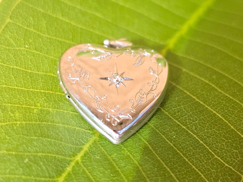 May include: A silver heart-shaped locket with a starburst design and floral accents. The locket is closed and sits on a green leaf. The locket has a shiny, reflective surface.
