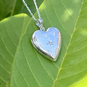 May include: A silver heart-shaped locket pendant on a delicate chain. The locket is engraved with floral and starburst designs, and a small gemstone is set in the center. The pendant rests on a large green leaf.