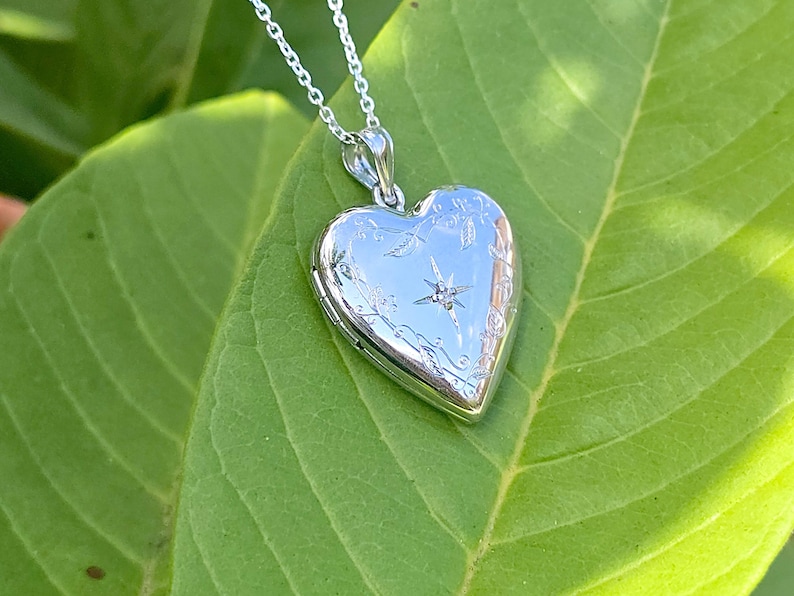 May include: A silver heart-shaped locket pendant on a delicate chain. The locket is engraved with floral and starburst designs, and a small gemstone is set in the center. The pendant rests on a large green leaf.
