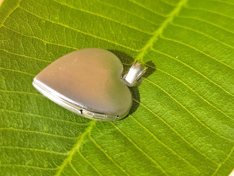 May include: A silver heart-shaped locket with a small loop for a chain, resting on a vibrant green leaf. The locket is closed, showing a smooth, reflective surface. The leaf's texture and veins are clearly visible.