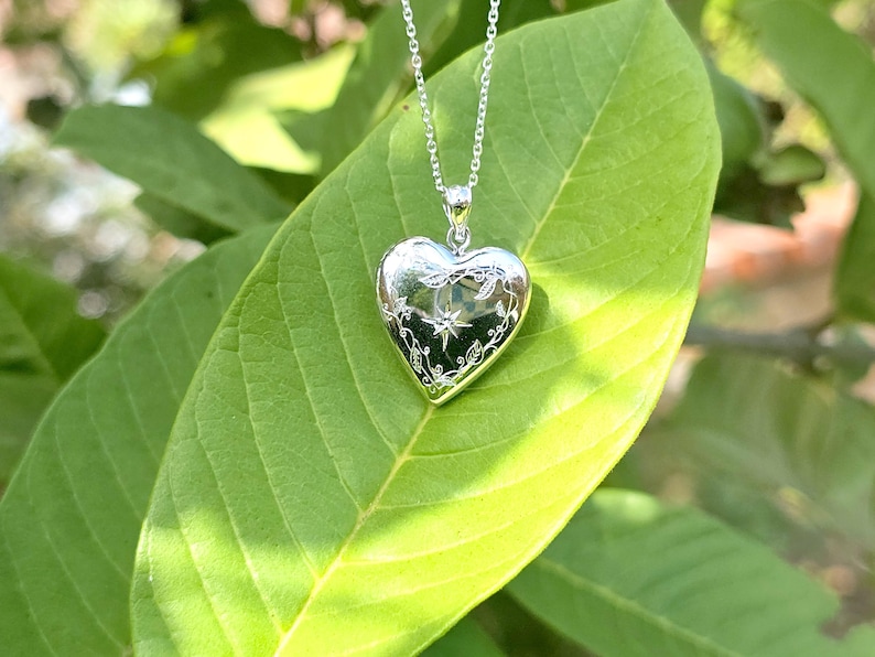 May include: A silver heart-shaped locket pendant with a delicate floral and star design, suspended from a fine chain. The locket rests on a large green leaf, with a blurred background of foliage.