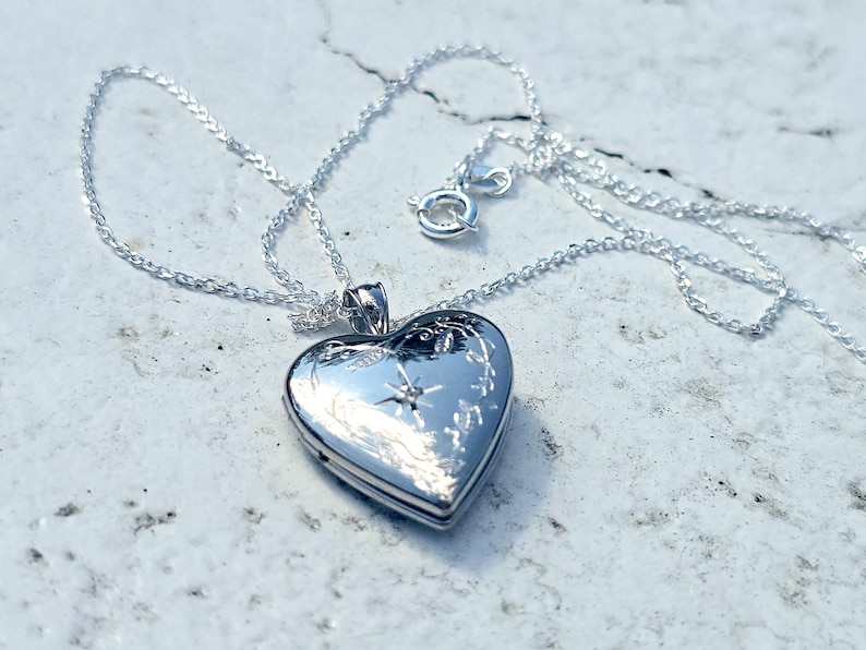 May include: A silver heart-shaped locket necklace. The locket is engraved with floral and starburst designs. The necklace chain and clasp are also silver. The locket is closed and reflects the light.